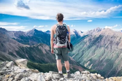 Mann mit dem Rücken zur Kamera. Er steht auf dem Berg und sieht über felsige Berggipfel und ins Tal.