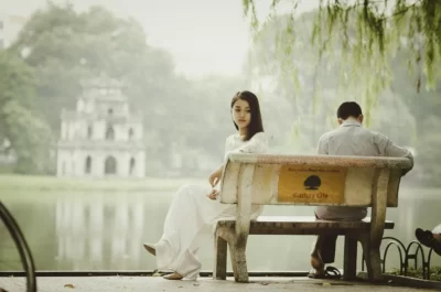 Asiatisches Paar im Park. Sie haben vor sich einen See, dahinter steht ein vierstöckiger Steinpavillon. Sie sitzen auf einer Steinbank. Der Mann sitzt rechts, blickt nach unten, ist mit dem Rücken zur Kamera. Die Frau sitzt links mit einem Abstand zu ihm am Rand der Bank und dreht sich zur Kamera. Ihr Blick wirkt traurig. Sie blickt leicht zum Boden.